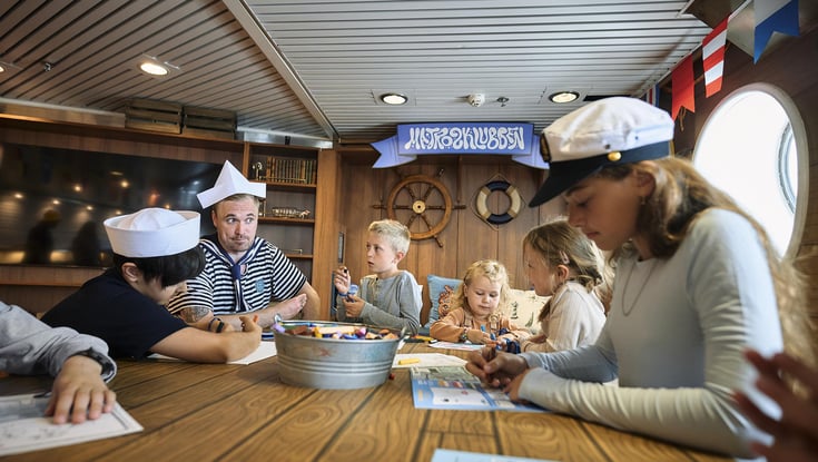 Group of people, including children and adults, engaged in an activity with paper and markers in the Matrosklubben.